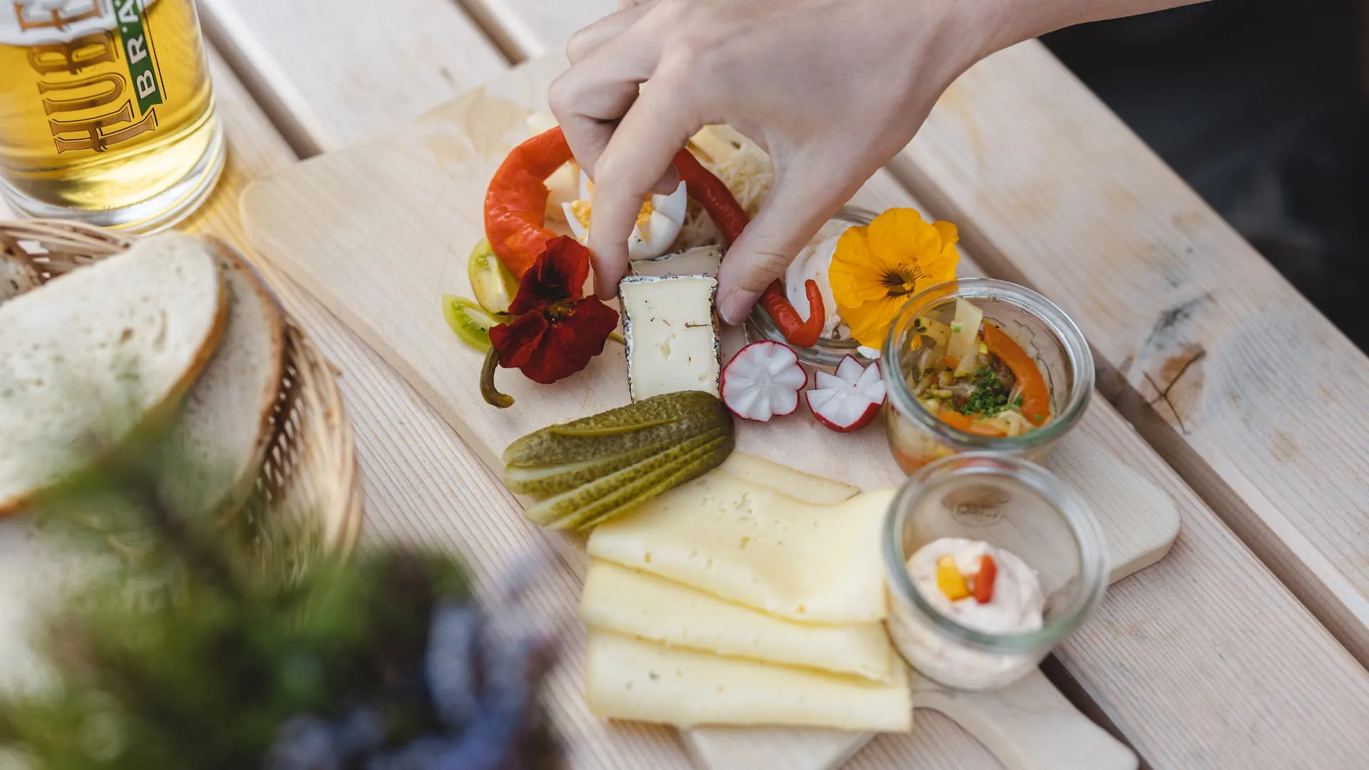 Teller mit Käse, Gemüse und Dips auf Holztisch, Hand legt Zutat hinzu