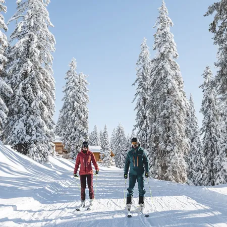 Zwei Skifahrer fahren eine verschneite Piste durch winterliche Bäume.